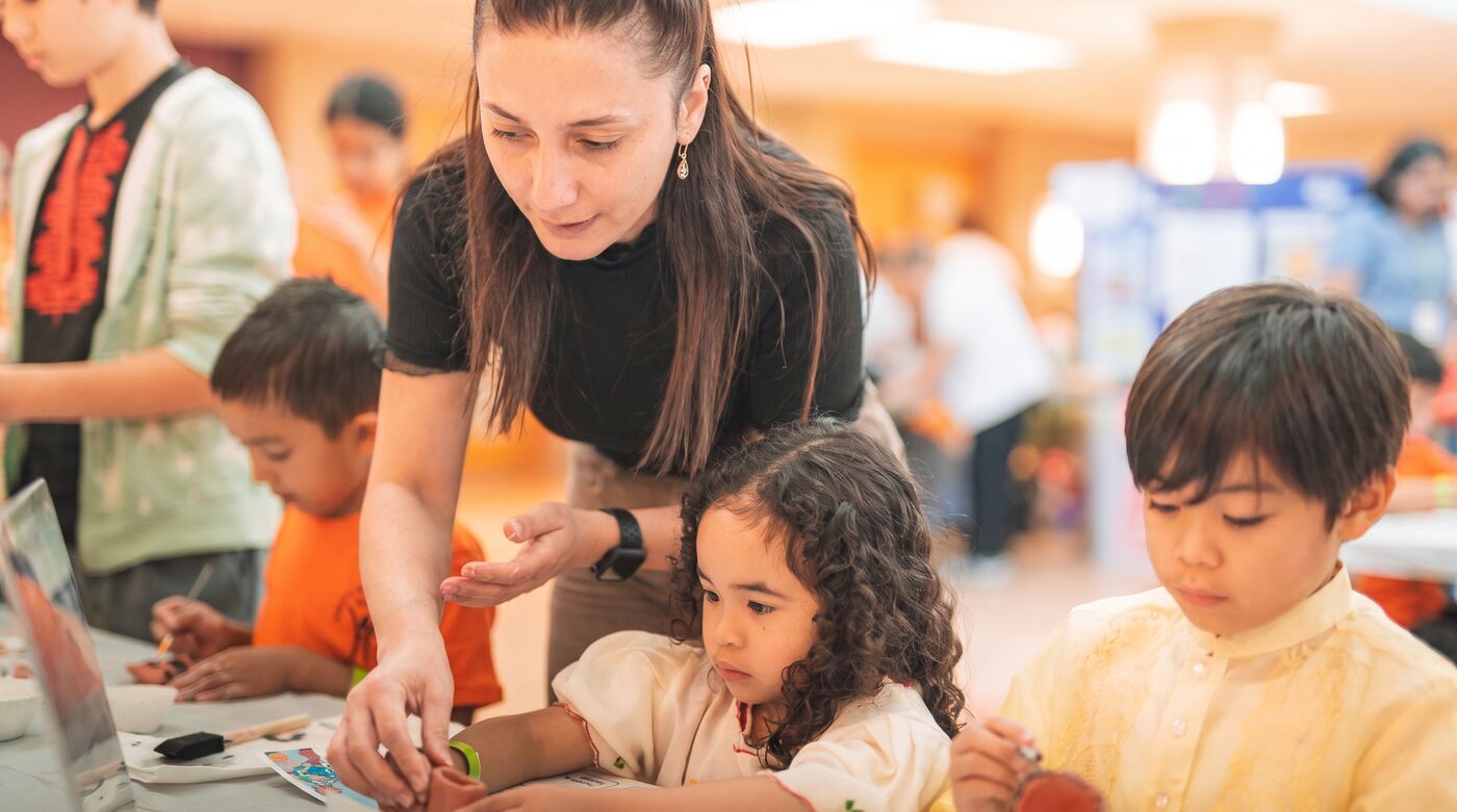 A teacher leaning across a table to guide two young children through a painting activity.